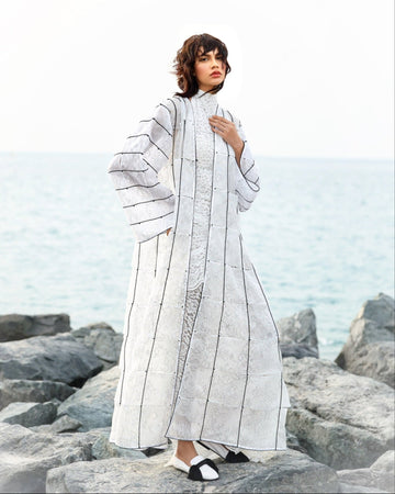 Woman wearing a long, light-colored coat with geometric patterns on a rocky beach.