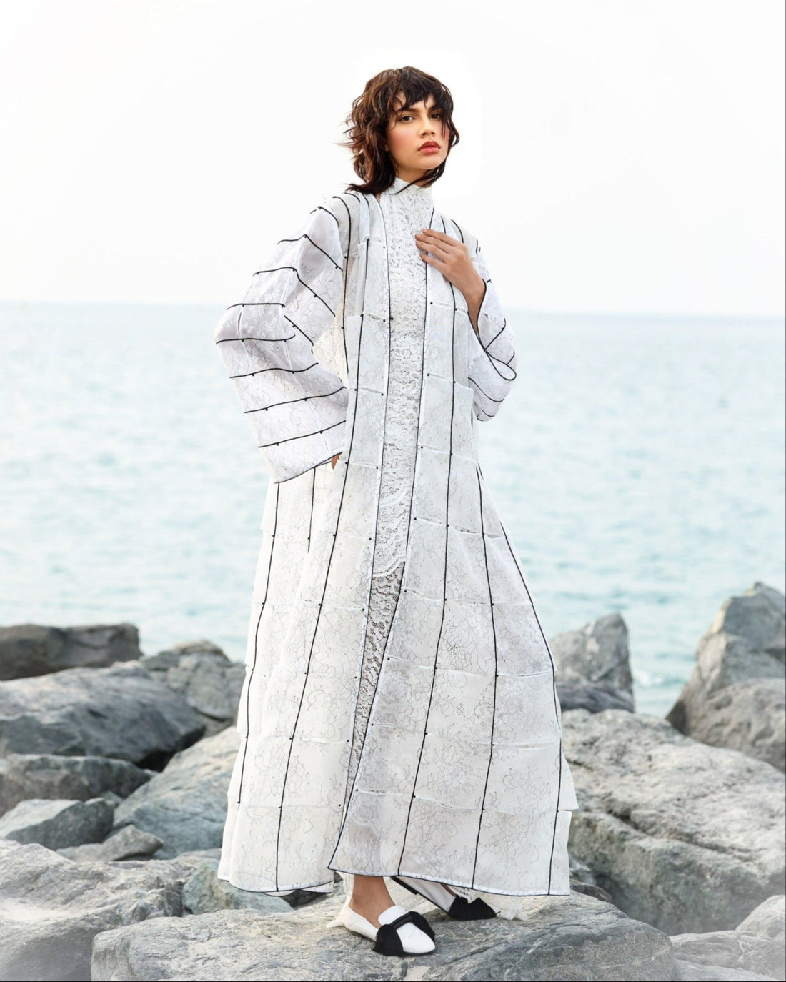 Woman wearing a long, light-colored coat with geometric patterns on a rocky beach.