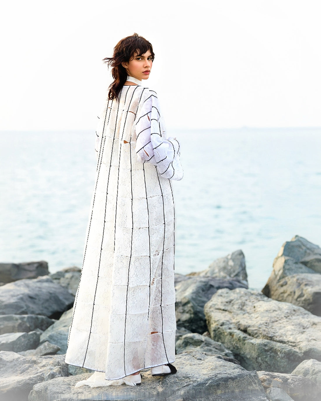 Woman wearing a long, light-colored coat with geometric patterns on a rocky beach.